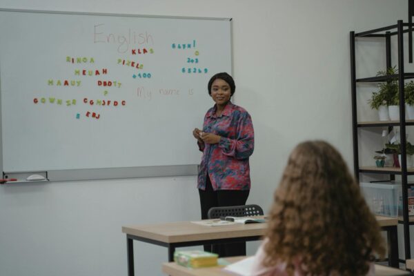 A teacher explaining English concepts to a student in a classroom setting with a whiteboard.